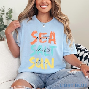 May include: A light blue short-sleeve t-shirt featuring a graphic design with the words SEA, SALT, and SUN printed in large, brush-stroke style letters in orange, teal, and yellow, with the words water, shells, and shine in black script.