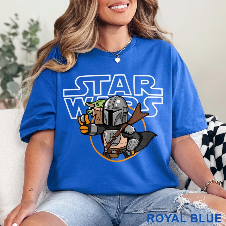 May include: Royal blue t-shirt featuring a Star Wars graphic. The design includes the Star Wars logo and an illustration of The Mandalorian with Grogu. The shirt is a vibrant blue and has the words "ROYAL BLUE" printed at the bottom.
