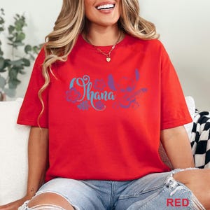 May include: Red t-shirt with the word "Ohana" in blue script, accompanied by a purple and blue graphic of a character and flowers. The word "RED" is printed in the bottom right corner of the image.