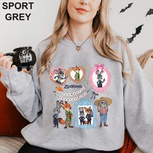 Op de afbeelding: Sportgrijs sweatshirt met Zootopia-karakterillustraties in frames. De sweatshirt heeft een ronde hals en lange mouwen. De tekst "Zootopia" en "welcome" zijn ook zichtbaar. Een zwarte mok met de tekst "Witches Brew" wordt in de afbeelding vastgehouden.