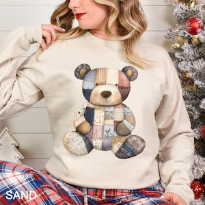 May include: A sand-colored sweatshirt featuring a patchwork teddy bear design. The bear has a brown nose, ears, and paws, with a body composed of various fabric patterns in shades of blue, pink, and beige. The word "SAND" is printed at the bottom.