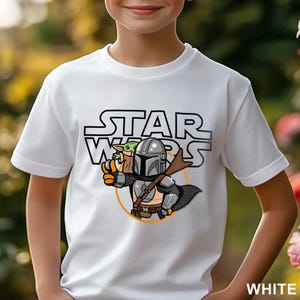 May include: White t-shirt featuring a Star Wars graphic. The design includes the Star Wars logo, a Mandalorian character, and Grogu. The Mandalorian is holding Grogu, with an orange circle around them. The word "WHITE" is in the bottom right corner.