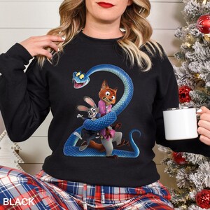 Puede incluir: Sudadera negra con una ilustraci&oacute;n de dibujos animados de una serpiente azul enrollada alrededor de un zorro y un conejo. El zorro lleva una camisa rosa y el conejo un traje. La palabra "BLACK" est&aacute; impresa en la parte inferior.