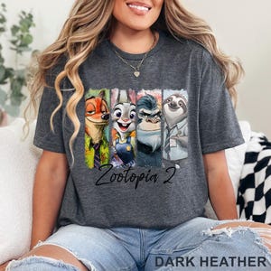 May include: Dark grey t-shirt with a Zootopia 2 design. The graphic shows Nick Wilde, Judy Hopps, a gorilla, and Flash the sloth. The words "Zootopia 2" are written in cursive script below the characters.