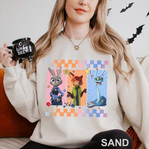 May include: Sand-coloured sweatshirt featuring three cartoon characters from Zootopia: Judy Hopps, Nick Wilde, and a snake. The design includes a checkered pattern in orange, pink, and blue. The sweatshirt is a comfortable, casual style.
