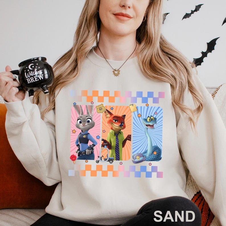 May include: Sand-coloured sweatshirt featuring three cartoon characters from Zootopia: Judy Hopps, Nick Wilde, and a snake. The design includes a checkered pattern in orange, pink, and blue. The sweatshirt is a comfortable, casual style.