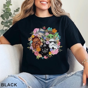 May include: Black t-shirt featuring a floral arrangement of Star Wars characters, including Chewbacca, Darth Vader, and a Stormtrooper. The design also includes R2-D2 and Grogu. The word "BLACK" is printed at the bottom.