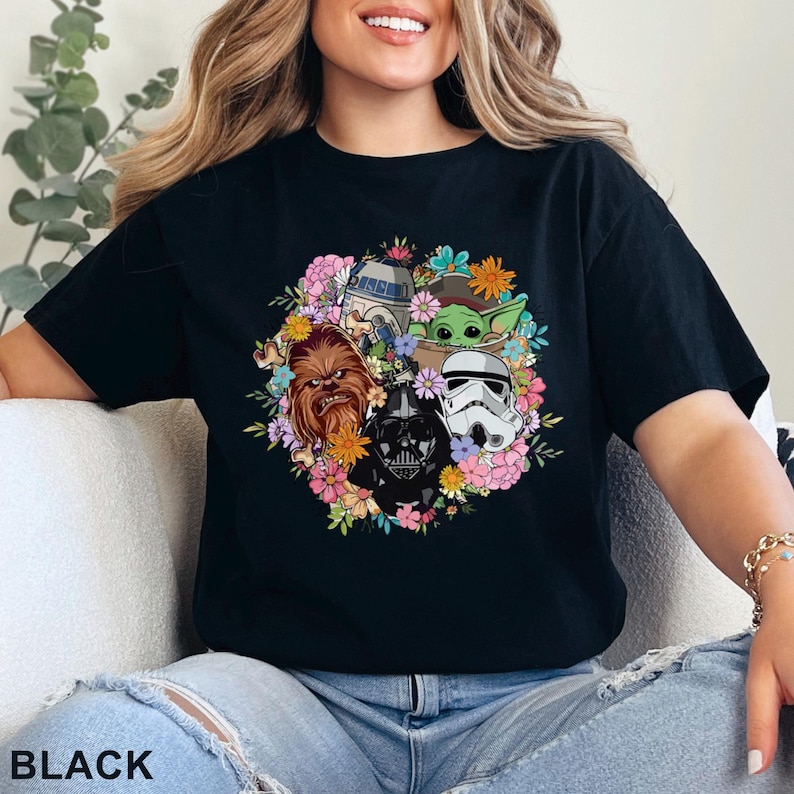 May include: Black t-shirt featuring a floral arrangement of Star Wars characters, including Chewbacca, Darth Vader, and a Stormtrooper. The design also includes R2-D2 and Grogu. The word "BLACK" is printed at the bottom.