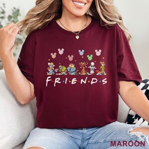 May include: Maroon t-shirt with cartoon characters and the word "FRIENDS" in white. Mickey Mouse head balloons in various colours float above the characters. The shirt is a solid maroon colour.
