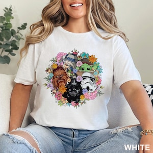 May include: White t-shirt featuring a colorful floral design with Star Wars characters, including Chewbacca, Darth Vader, a Stormtrooper, R2-D2, and Grogu. The word "WHITE" is printed at the bottom of the shirt.