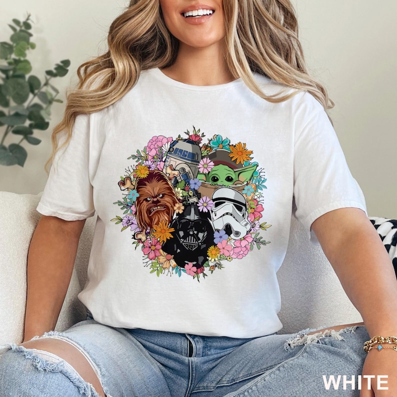 May include: White t-shirt featuring a colorful floral design with Star Wars characters, including Chewbacca, Darth Vader, a Stormtrooper, R2-D2, and Grogu. The word "WHITE" is printed at the bottom of the shirt.