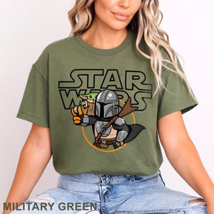 May include: Military green t-shirt featuring a cartoon illustration of The Mandalorian and Grogu from Star Wars. The design includes the Star Wars logo in black. The shirt is a comfortable, casual style.