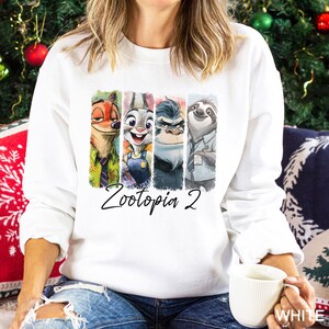 Op de afbeelding: Witte sweatshirt met een kleurrijke afbeelding van personages uit Zootopia 2. Het ontwerp bevat Nick Wilde, Judy Hopps, Chief Bogo en Flash. De tekst "Zootopia 2" is in cursieve letters onder de personages geschreven. Het woord "WHITE" staat rechtsonder afgedrukt.