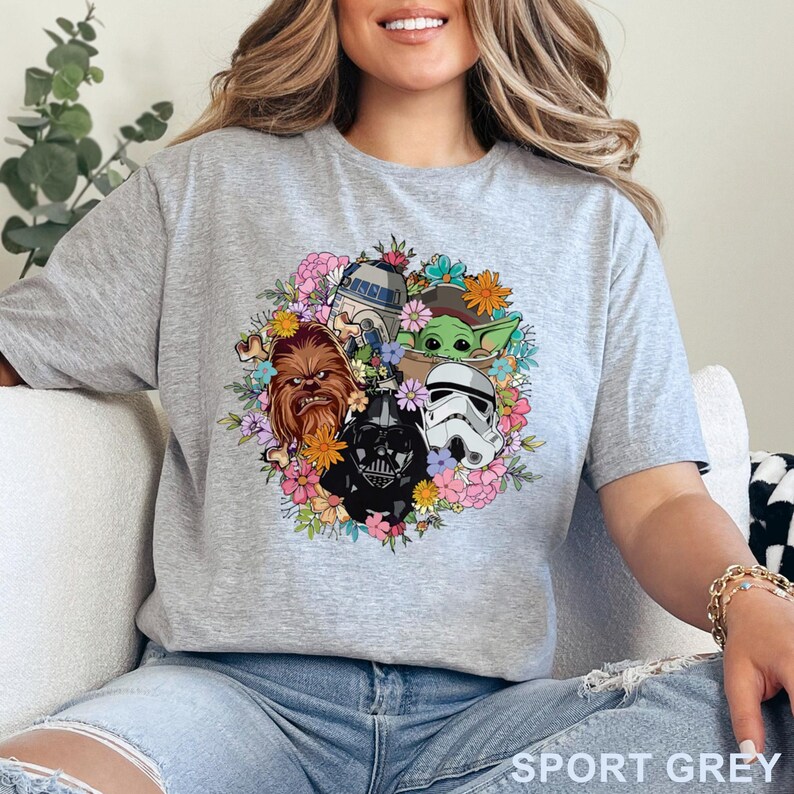 May include: Sport grey t-shirt featuring a floral wreath design with Star Wars characters including Chewbacca, Darth Vader, a Stormtrooper, R2-D2, and Grogu. The shirt has short sleeves and the text "SPORT GREY" is printed at the bottom.