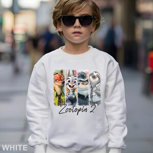 Op de afbeelding: Witte sweatshirt met een kleurrijk ontwerp met de personages uit Zootopia 2, waaronder Nick Wilde, Judy Hopps, Chief Bogo en Flash. De tekst "Zootopia 2" staat eronder gedrukt. De sweatshirt wordt gedragen door een persoon met een zonnebril.