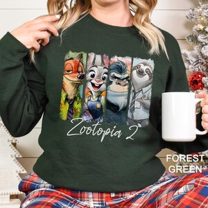 Op de afbeelding: Boszwarte sweatshirt met personages uit Zootopia. Het ontwerp bevat Nick Wilde, Judy Hopps, Chief Bogo en Flash. De tekst "Zootopia 2" is in witte letters geschreven. De woorden "Forest Green" staan eronder afgedrukt.