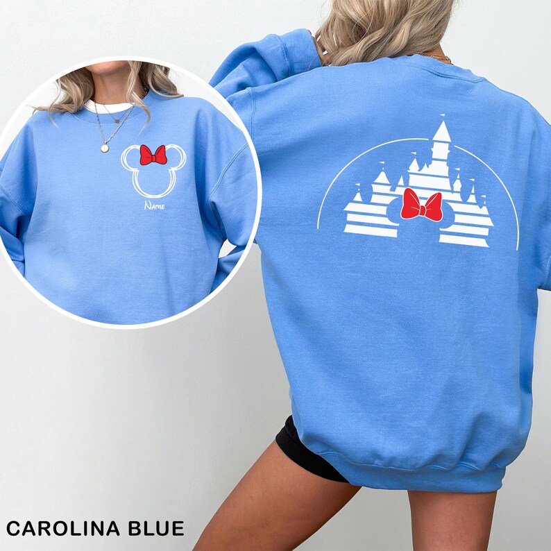 Pu&ograve; includere: Felpa blu Carolina con un contorno bianco di Minnie Mouse e un fiocco rosso, con il nome "Name". Il retro mostra un disegno di castello bianco e un fiocco rosso. Il testo "CAROLINA BLUE" &egrave; nella parte inferiore dell'immagine.