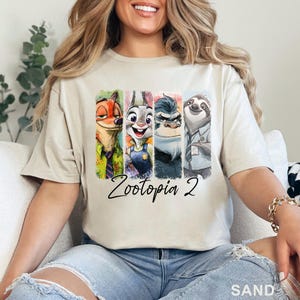 May include: Sand-coloured t-shirt with a vibrant graphic of Zootopia 2 characters. The design showcases Nick Wilde, Judy Hopps, Duke Weaselton, Chief Bogo, and Flash. The words "Zootopia 2" are written in a cursive font beneath the characters.