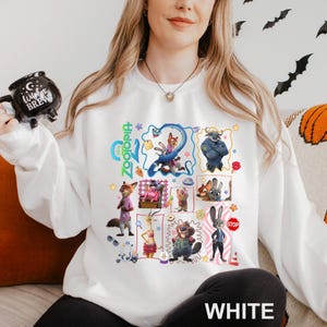 May include: White crewneck sweatshirt featuring a colorful graphic of characters from the animated movie Zootopia. The design includes Judy Hopps, Nick Wilde, and other characters. The word "Zootopia" is written in green. The word "WHITE" is printed at the bottom.