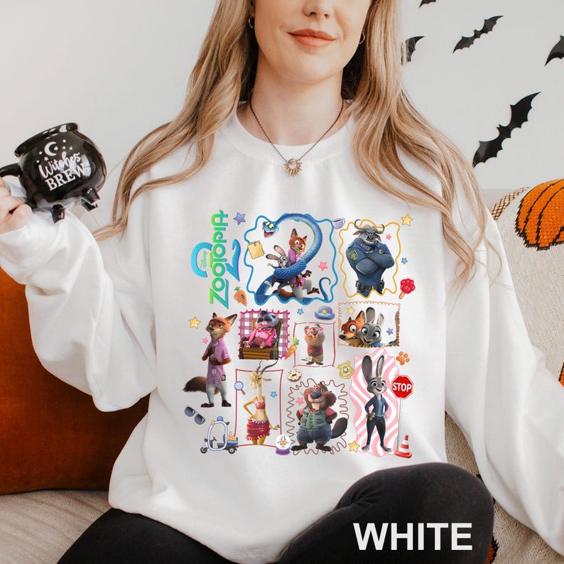 May include: White crewneck sweatshirt featuring a colorful graphic of characters from the animated movie Zootopia. The design includes Judy Hopps, Nick Wilde, and other characters. The word "Zootopia" is written in green. The word "WHITE" is printed at the bottom.