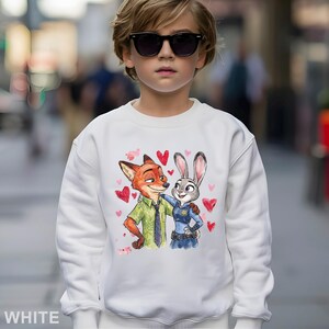 May include: White sweatshirt featuring a colorful illustration of Nick Wilde and Judy Hopps from Zootopia, surrounded by pink hearts. The sweatshirt has a crew neck and long sleeves. The word "WHITE" is printed at the bottom.