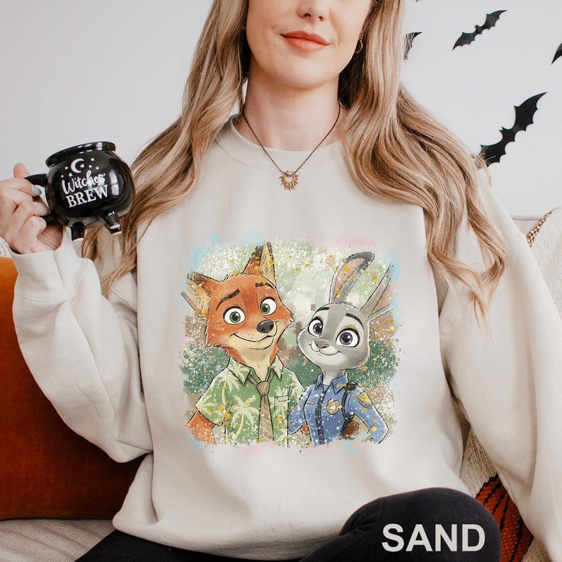 Puede incluir: Una sudadera de color arena con un gr&aacute;fico colorido de Nick Wilde y Judy Hopps de Zootopia. Nick lleva una camisa verde y Judy un uniforme de polic&iacute;a. La sudadera es de estilo casual.