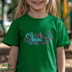 May include: Irish green t-shirt featuring the word "Ohana" in a stylized font with floral accents and a cartoon character. The design is in shades of blue and purple. The shirt is a short-sleeved crew neck.