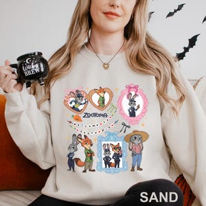 Op de afbeelding: Zandkleurige sweatshirt met kleurrijke cartoonafbeeldingen van personages uit de film Zootopia. Het ontwerp bevat ingelijste portretten van de personages, het woord "Zootopia" en de zin "welcome to Zootopia".