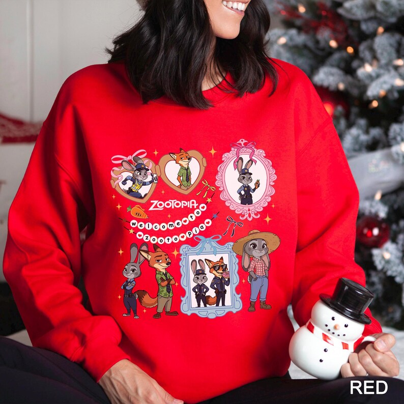 Op de afbeelding: Een rode sweatshirt met een ontwerp met personages uit "Zootopia". Het ontwerp bevat ingelijste portretten, harten en het woord "Zootopia". Een sneeuwpopfiguur wordt in de rechter benedenhoek vastgehouden.
