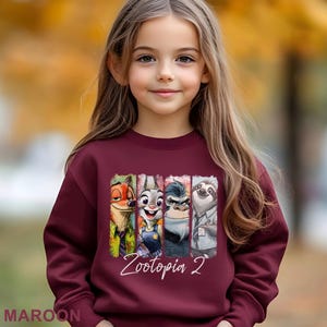 Op de afbeelding: Een bordeauxrode sweatshirt met een kleurrijke afbeelding van personages uit Zootopia 2. De afbeelding bevat Nick Wilde, Judy Hopps, een gorilla en Flash de luiaard. De tekst "Zootopia 2" staat in witte letters. Het woord "MAROON" staat onderaan.