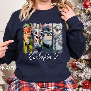 Op de afbeelding: Marineblauwe sweatshirt met een kleurrijke afbeelding van personages uit de film "Zootopia 2". Het ontwerp toont Nick Wilde, Judy Hopps, Chief Bogo en Flash. De woorden "Zootopia 2" staan in witte letters eronder.