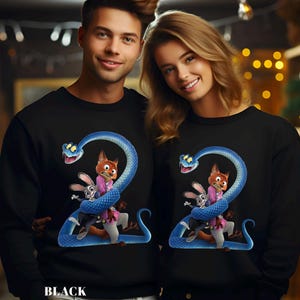 Puede incluir: Sudaderas negras con un dise&ntilde;o de serpiente azul y personajes de dibujos animados. El dise&ntilde;o incluye un zorro, un conejo y una serpiente que forman el n&uacute;mero "2". La palabra "BLACK" est&aacute; impresa en la parte inferior.