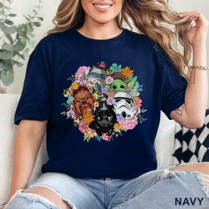 May include: Navy blue t-shirt featuring a floral arrangement with Star Wars characters including Darth Vader, Chewbacca, and Baby Yoda. The design incorporates colorful flowers and the text "NAVY" at the bottom.