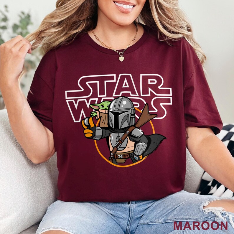 May include: Maroon t-shirt featuring a cartoon illustration of The Mandalorian and Grogu from Star Wars. The Star Wars logo is printed in white above the characters. The word "MAROON" is printed in white at the bottom of the shirt.
