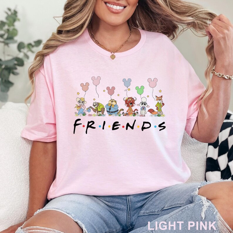May include: Pale pink t-shirt with the word "FRIENDS" in a colourful font, and cartoon characters holding balloon bouquets. The shirt is a light pink colour.