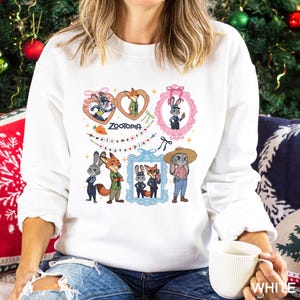 Op de afbeelding: Witte sweatshirt met kleurrijke cartoonfiguren en de tekst "Zootopia". Het ontwerp bevat ingelijste afbeeldingen van een konijn en een vos, samen met andere personages en decoratieve elementen. De sweatshirt heeft een ronde hals.