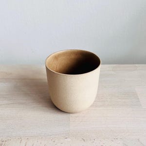 Puede incluir: Una taza de cerámica beige con un exterior moteado y un interior liso y marrón. La taza tiene una forma redondeada, ligeramente cónica y se apoya sobre una superficie de madera de color claro. La taza está vacía.