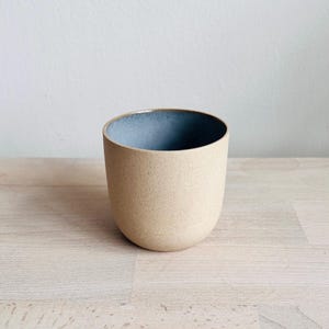 Puede incluir: Una taza de cerámica beige con una textura moteada y una base redondeada. El borde interior es de color azul contrastante. La taza se muestra sobre una superficie de madera clara.