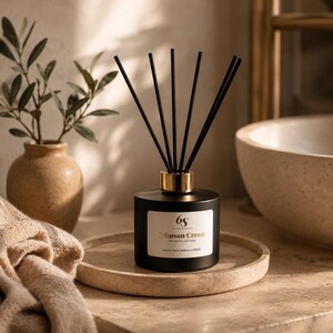 May include: A black reed diffuser with a gold collar and black reeds, labelled "Crimson Cream" on a white label. The diffuser sits on a round, beige tray. A beige vase with greenery and a beige towel are also in the image.