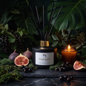 May include: A matte black reed diffuser bottle with a gold collar and black reeds. The white label reads 6s Sixth Scents Green Haven Green Fig | Ripe Cassis Luxury Reed Diffuser | 200ml. A lit amber glass candle sits beside it.