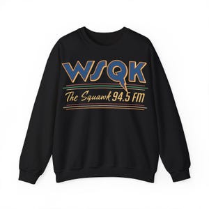 Peut inclure: Sweat-shirt noir avec l'inscription "WSQK The Squawk 94.5 FM" en bleu, or et rouge. Le design comprend un éclair. Le sweat-shirt a des manches longues et un col rond.