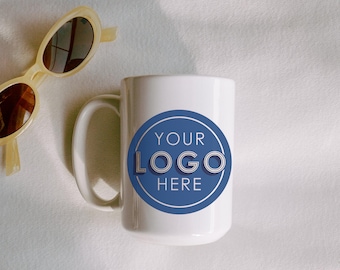 May include: White ceramic mug with a blue circular logo that reads "YOUR LOGO HERE." The mug has a curved handle and is next to a pair of cream-colored sunglasses. A disco ball and a "We Ship From USA" sticker are also visible.
