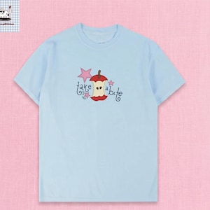 May include: Light blue t-shirt featuring a whimsical design of a red apple core with a bite taken out, a pink star, and the words "take a bite" in a playful font. The shirt is a short-sleeved crew neck.