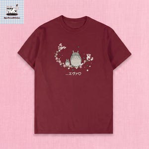 May include: Burgundy t-shirt featuring an embroidered design of two cartoon characters with a floral branch and Japanese text. The design includes a large and small character with a heart symbol. The shirt has short sleeves.