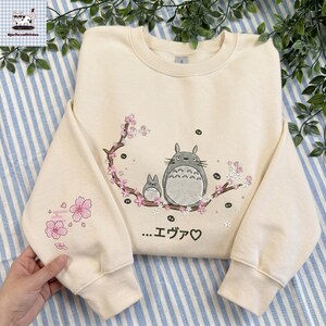 May include: Cream-colored sweatshirt featuring a Totoro design with a branch of pink cherry blossoms. The design includes two Totoro characters and Japanese text. The left sleeve has additional pink flower details.