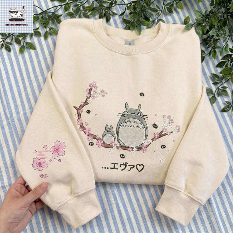 May include: Cream-colored sweatshirt featuring a Totoro design with a branch of pink cherry blossoms. The design includes two Totoro characters and Japanese text. The left sleeve has additional pink flower details.