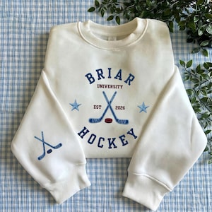 Puede incluir: Una sudadera blanca con bordados azules. Muestra "Briar University Hockey", palos de hockey cruzados, estrellas y el año "2026". La manga presenta un diseño de palo y disco de hockey.