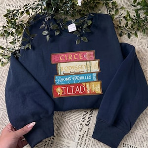 以下が含まれることがあります： 積み重ねられた本の刺繍デザインが施されたネイビーブルーのスウェットシャツ。「Circe」、「Odyssey」、「Song of Achilles」、「The Iliad」のタイトルが含まれています。スウェットシャツは長袖とクルーネックで、読書好きに最適です。