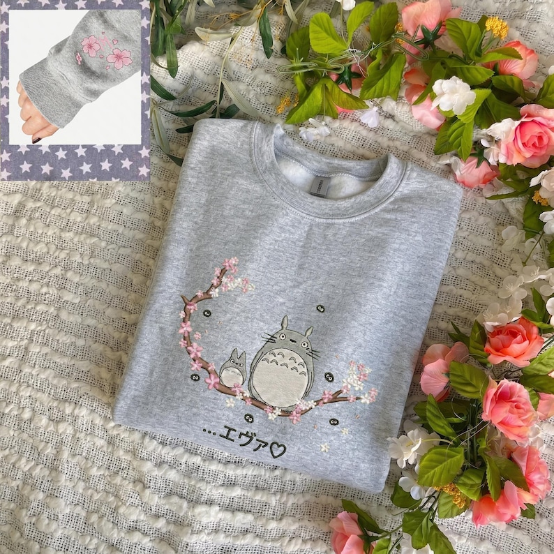 May include: A light gray sweatshirt featuring an embroidered design of two cartoon characters on a flowering branch. The design includes the Japanese text "...Eva♡". The sleeve cuff has pink cherry blossom embroidery.