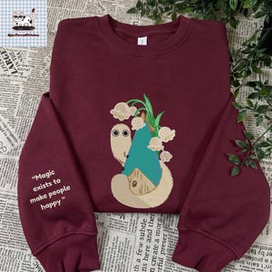 Op de afbeelding: Een bordeauxrood sweatshirt met ronde hals en een afbeelding van een beige wezen met grote ogen naast een huis met een blauw dak en lelietjes-van-dalen. Op de linkermouw staat de witte tekst: "Magic exists to make people happy."